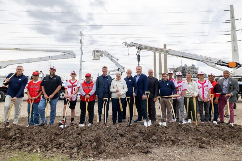 A group of people, including adults in business attire and youths in baseball uniforms, stand in a row outdoors holding shovels at a groundbreaking ceremony on the Salt Lake westside business district near North Temple Jordan River.
