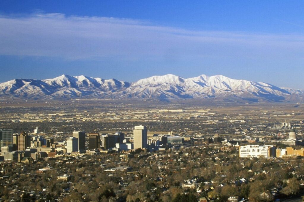 A cityscape featuring numerous buildings in the foreground and snow-capped mountains in the background, with the North Temple Jordan River area poised for growth amid talks of an MLB expansion Salt Lake City.
