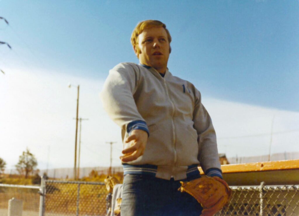 A man wearing a light jacket holds a baseball glove and stands near a chain-link fence on a sunny day in the Salt Lake westside business district.