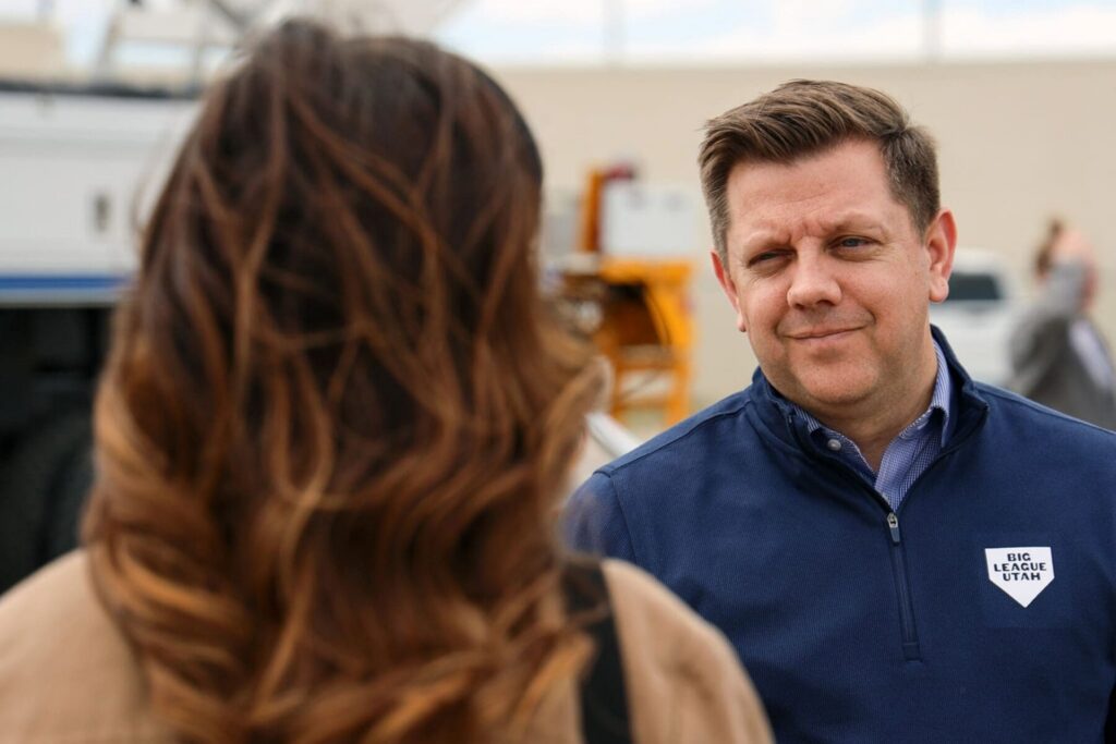 A man in a navy blue jacket speaks to a woman with long brown hair outdoors in the Salt Lake westside business district; industrial equipment is visible in the background.
