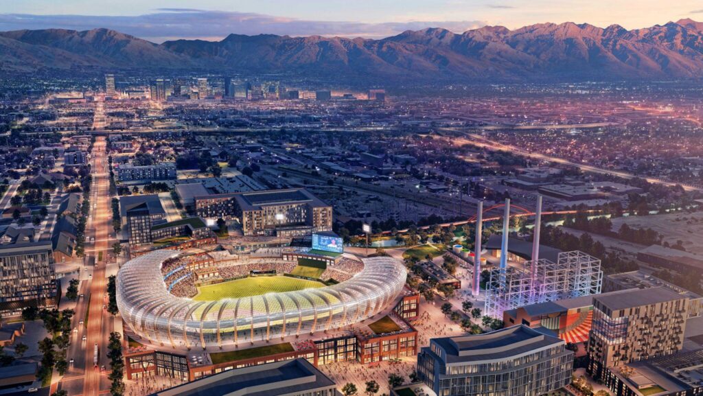 Aerial view of a modern sports stadium near the Salt Lake westside business district, with a cityscape and mountains in the background during dusk.