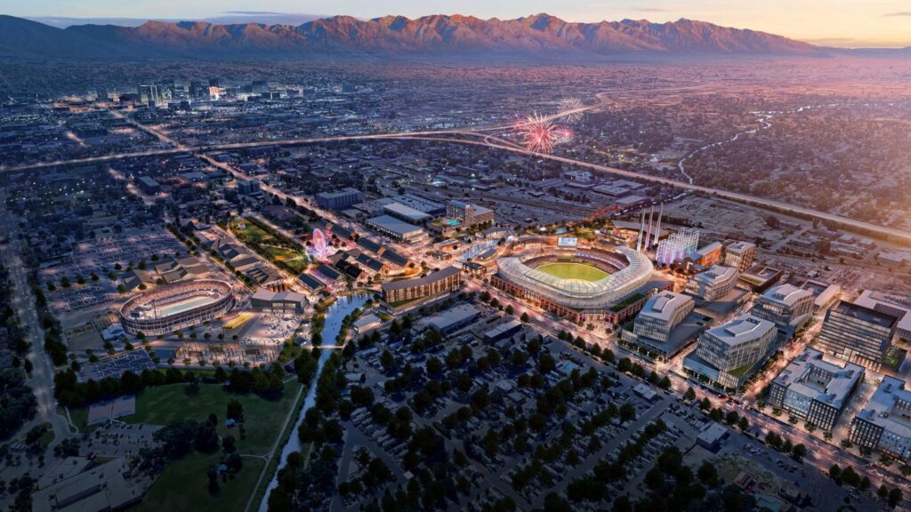 Aerial view of a city at sunset featuring sports stadiums, surrounding buildings, parking lots, and fireworks in the distance near the Salt Lake westside business district and North Temple Jordan River, with mountains framing the skyline.
