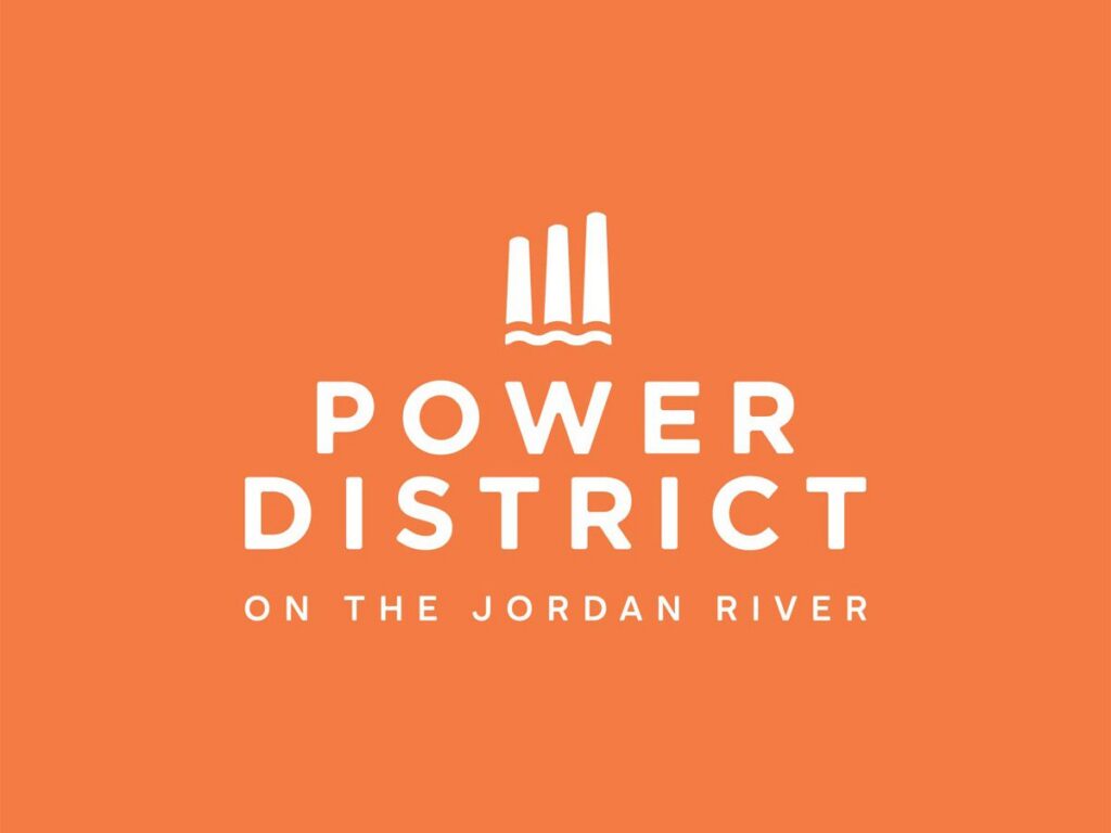 Orange background with a white logo above the text "POWER DISTRICT ON THE JORDAN RIVER." The logo features three vertical bars above two wavy lines, representing Salt Lake’s westside business district and an all-electric mixed-use development.