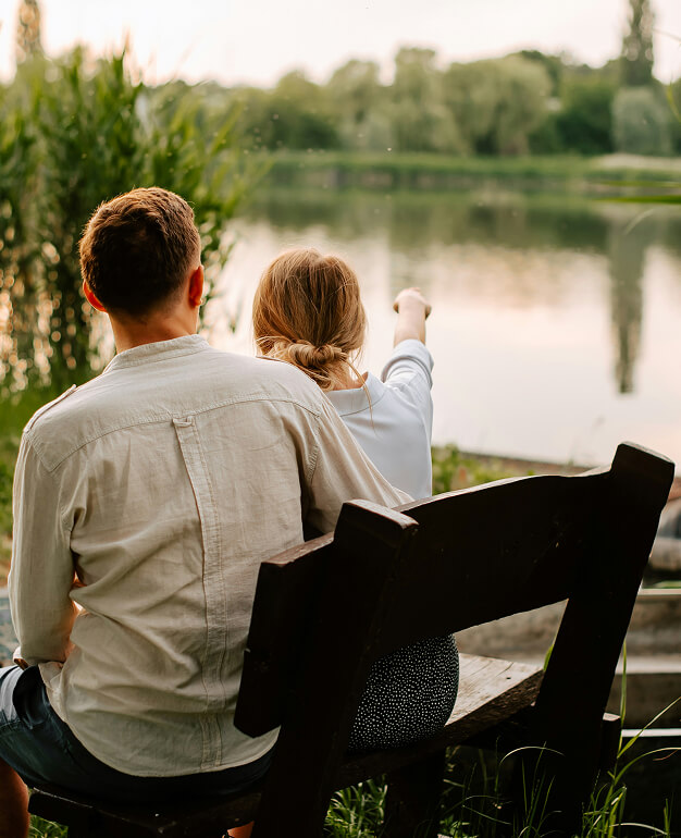 Two people sit on a bench by a lake near the Salt Lake westside business district, with one person pointing toward the water. Trees and foliage are visible in the background.