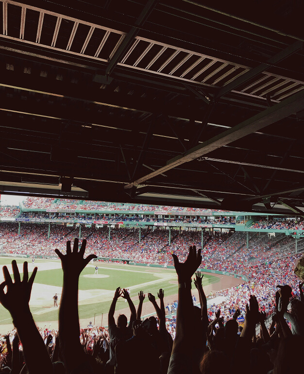 Fans with raised arms cheer at a crowded baseball stadium in the Salt Lake westside business district, viewed from under a shaded section, with the field and players visible in the background.