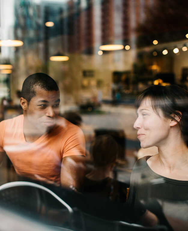 Two people sit at a table in a cafe, talking and smiling, seen through a window with reflections of the all-electric mixed-use development along North Temple Jordan River and the street lights outside.