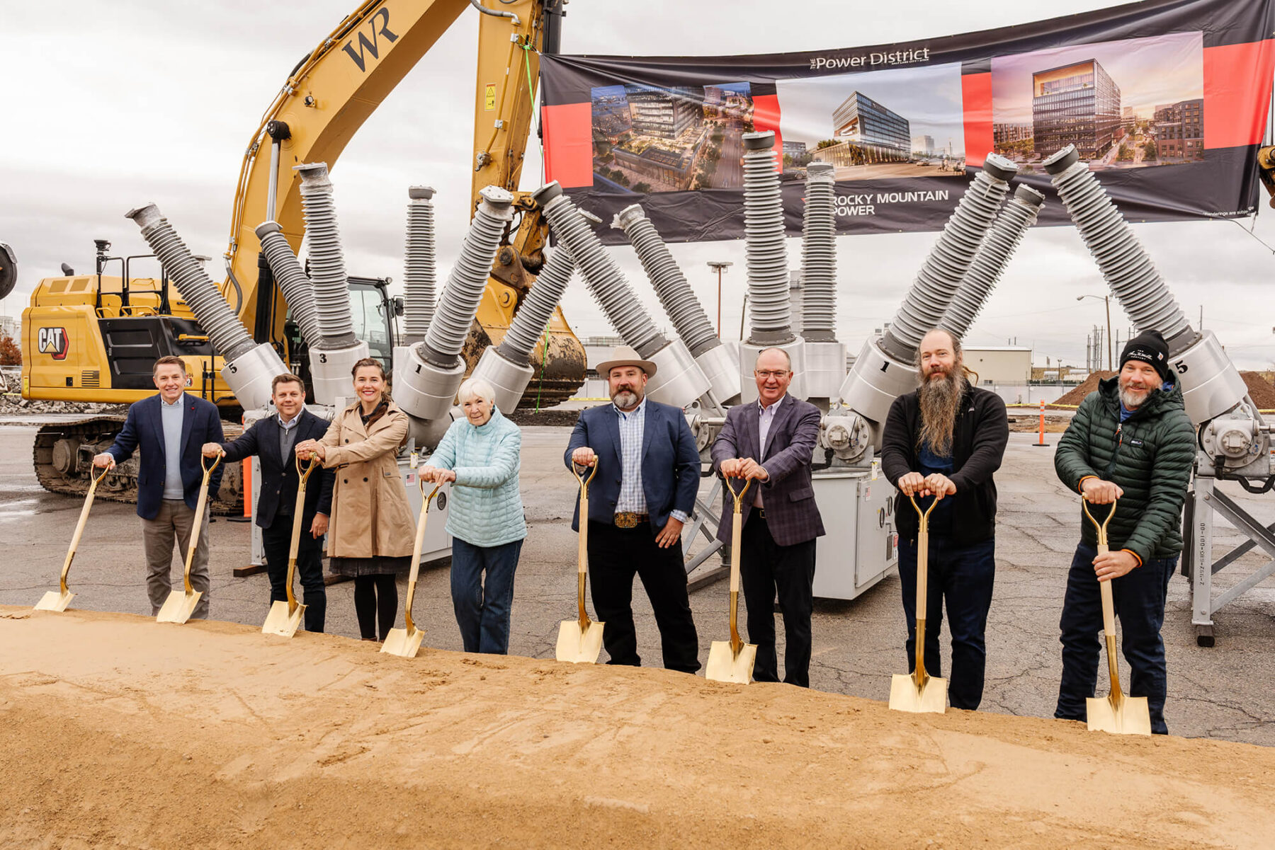 Ocho personas vestidas de traje posan con palas doradas en una ceremonia de inauguración en el distrito comercial oeste de Salt Lake, frente a equipos de construcción y una pancarta bajo un cielo nublado.
