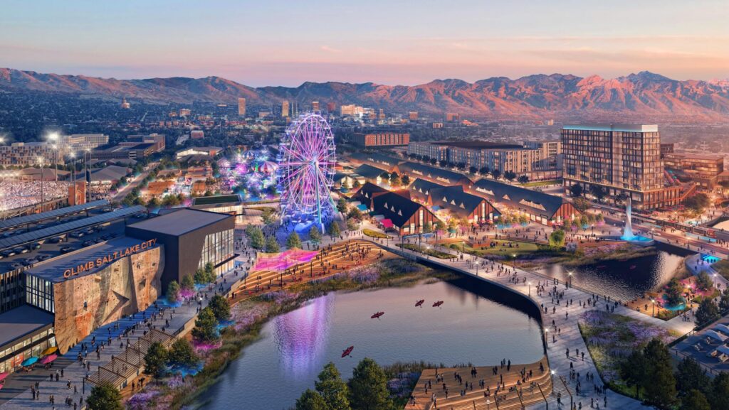 Aerial view of a vibrant entertainment district at sunset with a Ferris wheel, modern buildings, and people strolling along the North Temple Jordan River in Salt Lake City’s westside business district, all set against majestic mountain backdrops.