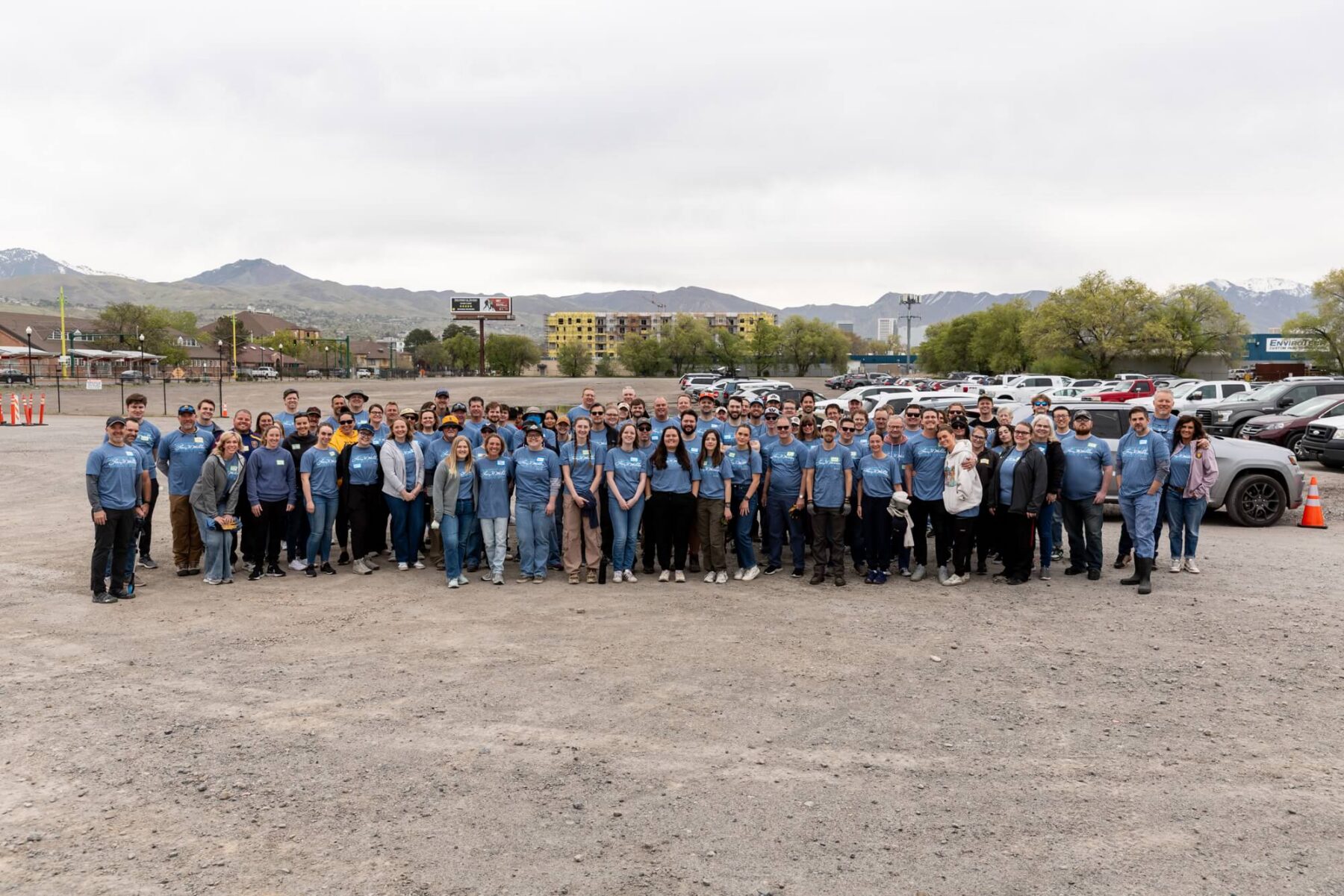 Un gran grupo de personas con camisetas azules posa junta en una zona de aparcamiento de grava cerca del río Jordan de North Temple, con montañas y coches aparcados al fondo.