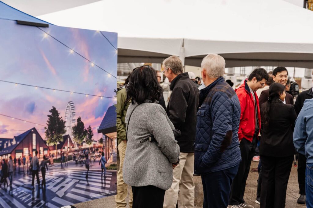 Un grupo de personas se encuentra cerca de un tablero de exhibición con una representación arquitectónica en un evento al aire libre bajo una gran carpa blanca, destacando el desarrollo listo para comenzar en Utah, cerca del río Jordan en North Temple.