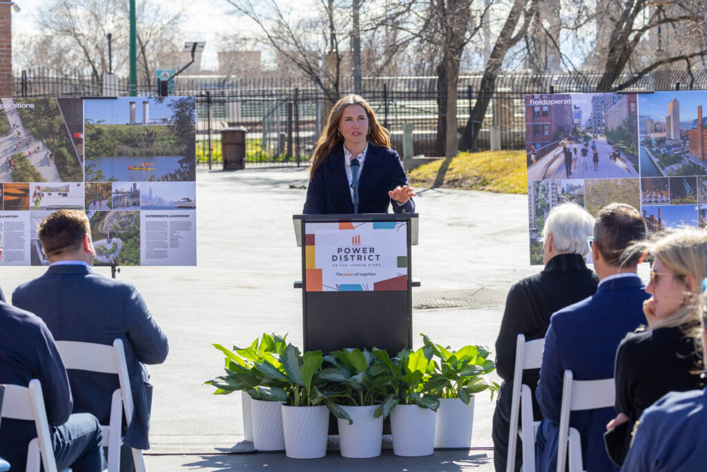 Una mujer habla en un podio al aire libre con la etiqueta "Power District" frente a asistentes sentados, con tableros de exhibición de proyectos y plantas alrededor del escenario en el distrito de negocios del oeste de Salt Lake.