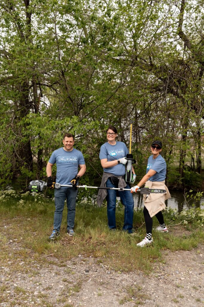 Tres personas están afuera frente a árboles, vistiendo camisas azules a juego y portando herramientas de jardinería, pareciendo participar en una limpieza comunitaria o evento de trabajo al aire libre de un distrito comercial del oeste de Salt Lake.