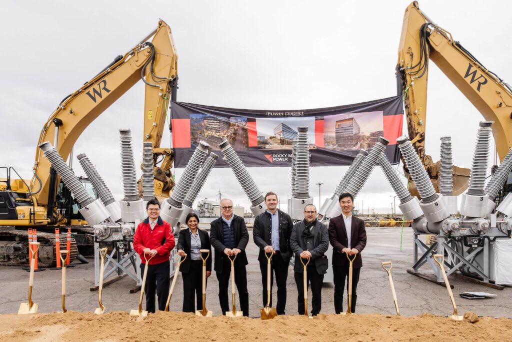 Seis personas vestidas de traje se paran con palas en una ceremonia de inauguración para un desarrollo listo para comenzar en el distrito de negocios del oeste de Salt Lake en Utah, con equipo de construcción y un cartel que muestra representaciones del edificio detrás de ellas.