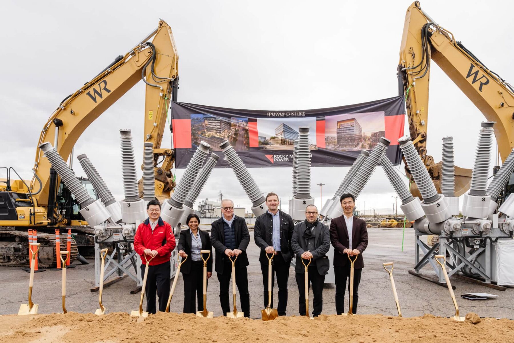 Seis personas vestidas de traje se paran con palas en una ceremonia de inauguración para un desarrollo listo para comenzar en el distrito de negocios del oeste de Salt Lake en Utah, con equipo de construcción y un cartel que muestra representaciones del edificio detrás de ellas.