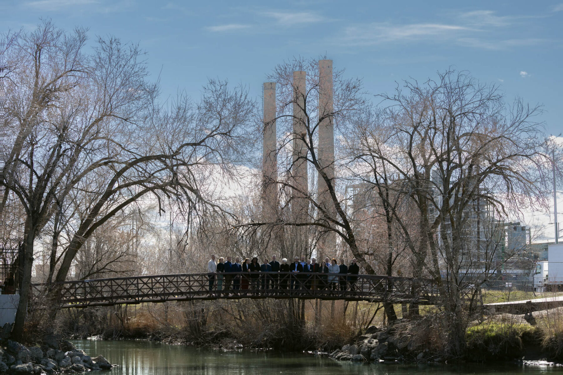 Un grupo de personas se para en un puente de madera sobre el río Jordán del Templo del Norte, con árboles sin hojas y chimeneas industriales visibles al fondo.
