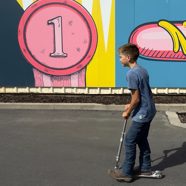 Niño andando en patineta sobre pavimento frente a un mural colorido que presenta un círculo rosa grande con el número 1 y parte de un zapato de caricatura, cerca del desarrollo de uso mixto totalmente eléctrico junto al río Jordan de North Temple.