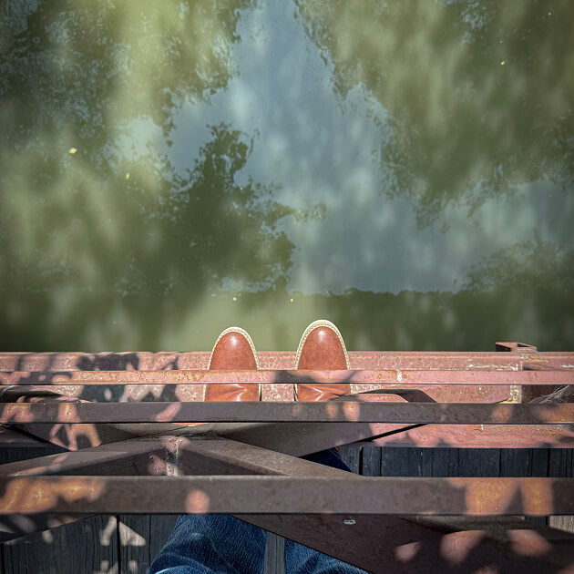 Persona con zapatos marrones y jeans azules parada en un puente de metal oxidado sobre el río Jordán, North Temple, mirando hacia el agua verdosa que fluye a través del distrito comercial del oeste de Salt Lake.