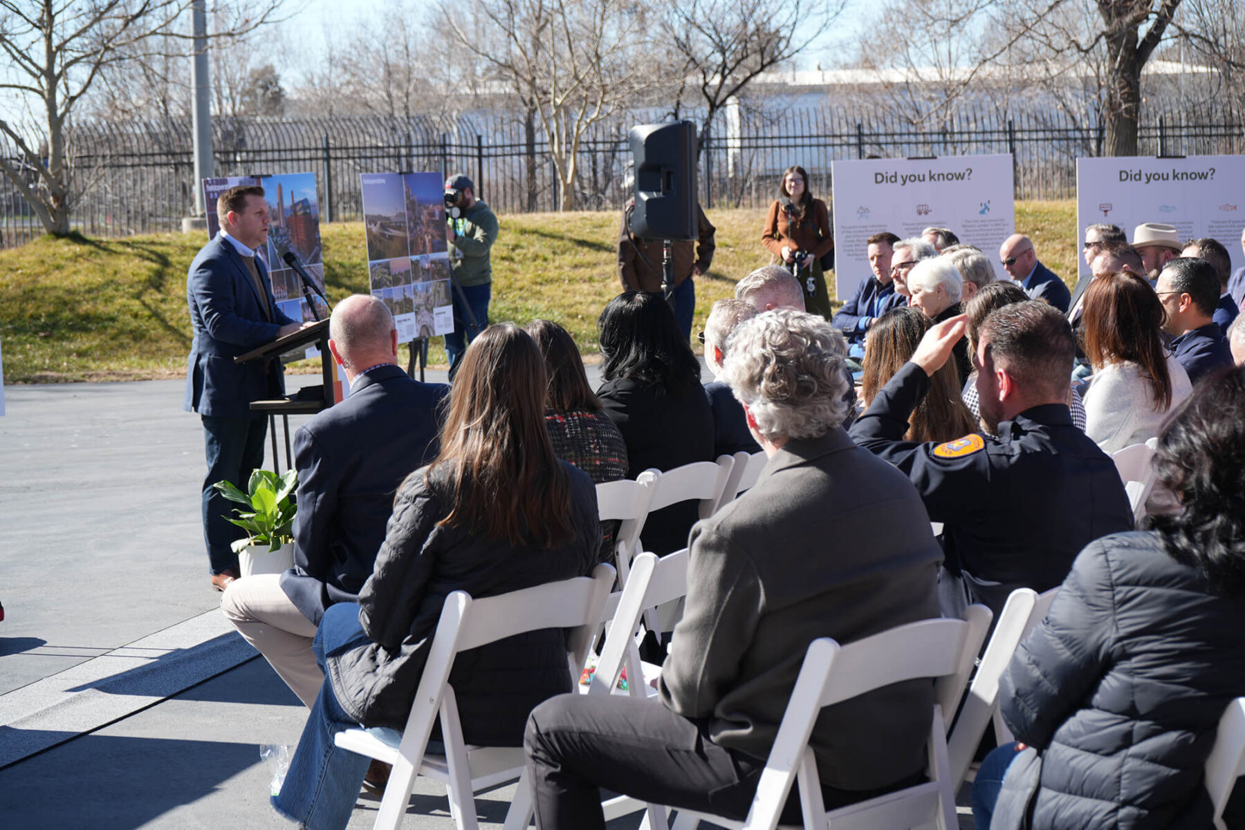 A man speaks at a podium to an audience seated outdoors near North Temple Jordan River; informational posters about MLB expansion Salt Lake City and a photographer are visible in the background.