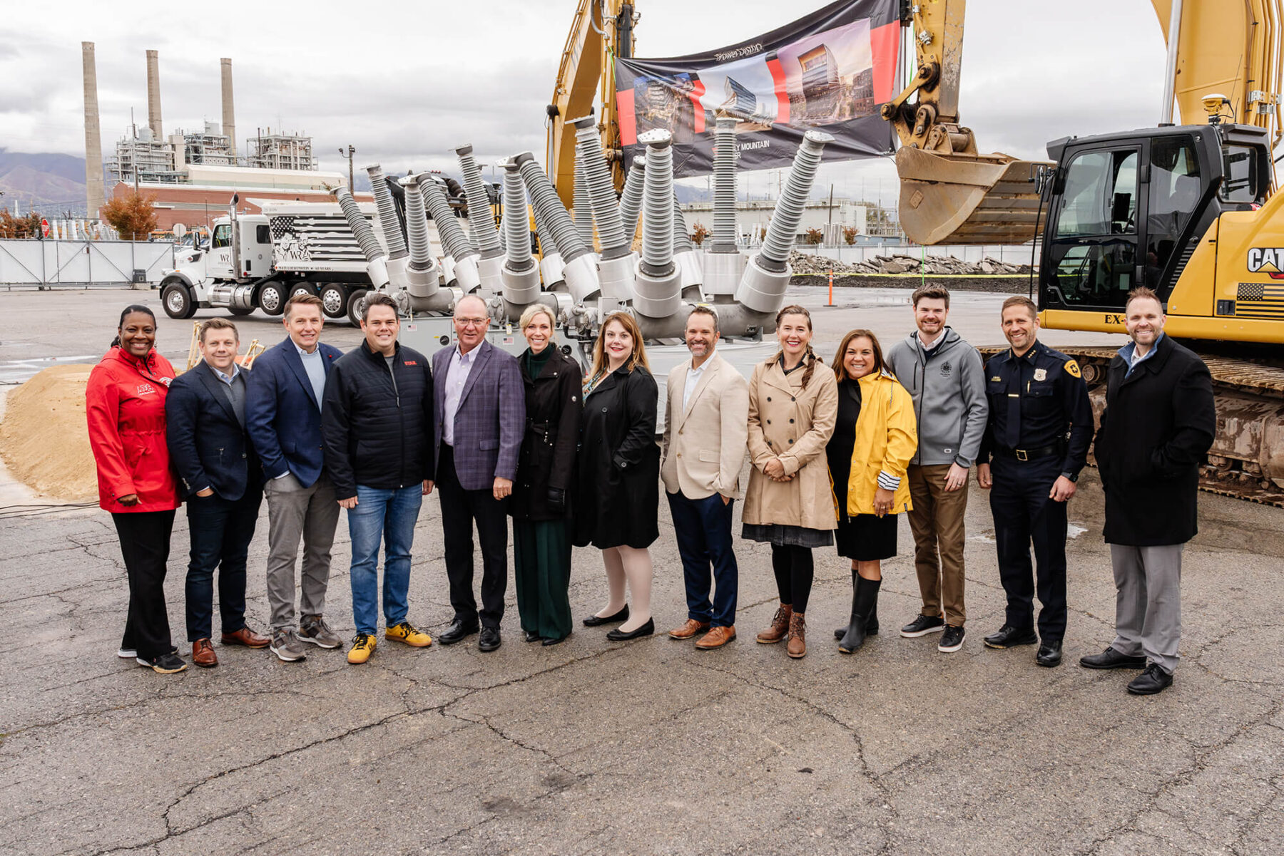 A group stands in front of construction equipment within the Salt Lake westside business district, posing for a photo on a cloudy day at the site of an all-electric mixed-use development.