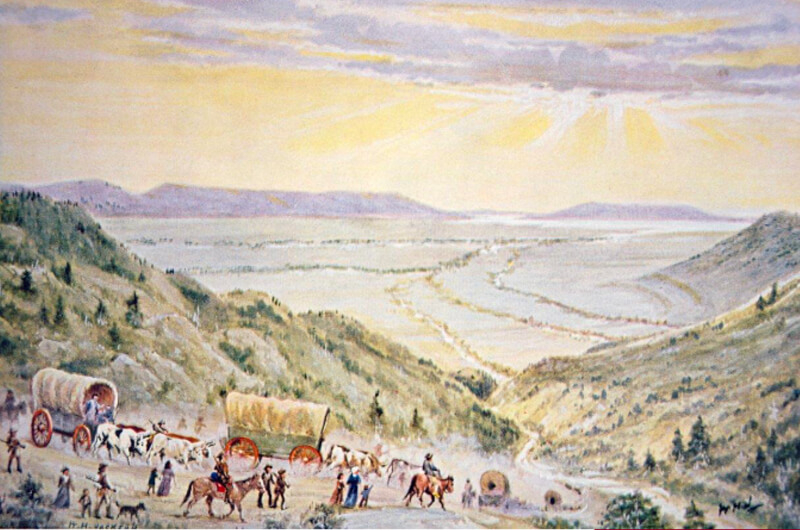 Un grupo de carretas cubiertas y pioneros descienden por un paso de montaña hacia un amplio valle con un lago en la distancia, cerca de donde el río Jordan del Templo del Norte se une al actual distrito de negocios del lado oeste de Salt Lake, bajo un cielo parcialmente nublado.