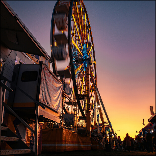 A Ferris wheel lit with colorful lights spins at a fairground during sunset, set against the backdrop of the North Temple Jordan River in Salt Lake’s westside business district, with silhouettes of people and booths visible.