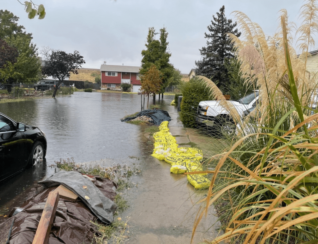 A residential street in the Salt Lake westside business district is flooded with water, sandbags line a driveway, and cars are parked nearby under overcast skies.