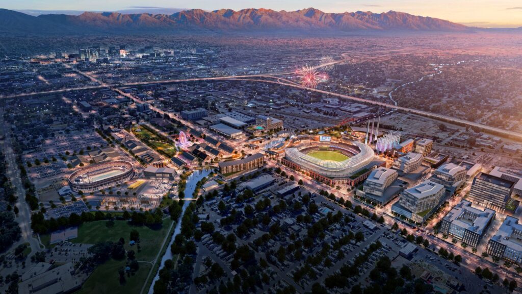 Vista aérea de una gran ciudad al atardecer con un complejo de estadios brillantemente iluminado —posible expansión de la MLB en Salt Lake City— fuegos artificiales en el cielo, montañas al fondo y el río Jordan de North Temple serpenteando junto a un desarrollo de uso mixto totalmente eléctrico.