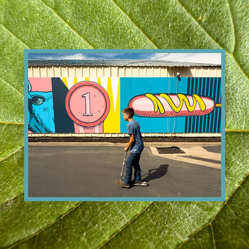 Un niño anda en patineta sobre el pavimento en el distrito de negocios del lado oeste de Salt Lake, pasando frente a un colorido mural de una moneda y un hot dog, con un borde texturizado de hojas verdes alrededor de la imagen.