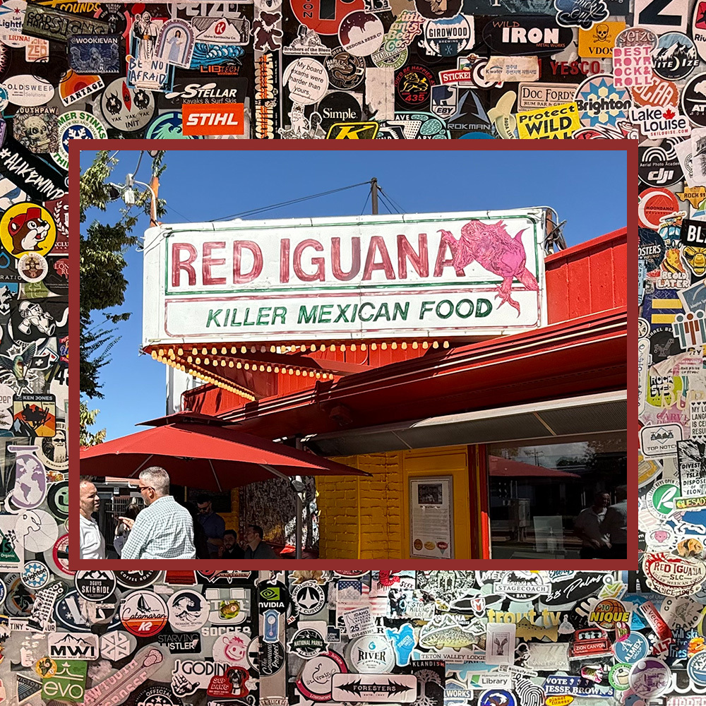 Letrero de “Red Iguana Killer Mexican Food” sobre un edificio rojo y amarillo en el vibrante distrito comercial del oeste de Salt Lake, enmarcado por coloridos adhesivos. Dos personas están paradas bajo una sombrilla roja.
