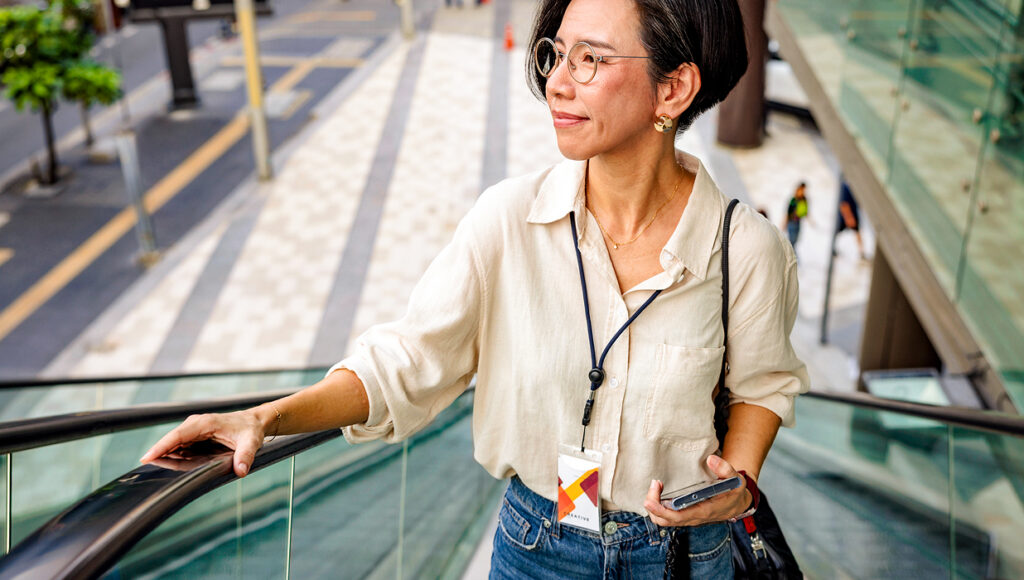 Una persona con pelo corto y gafas sube una escalera mecánica al aire libre, sosteniendo un teléfono inteligente y usando un cordón, con una calle y edificios —potencialmente parte de un desarrollo mixto totalmente eléctrico— de fondo.