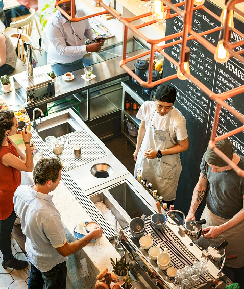 Vista aérea de una concurrida cafetería dentro de un desarrollo mixto totalmente eléctrico cerca del río Jordan de North Temple, con baristas preparando bebidas detrás del mostrador y clientes esperando o pidiendo cerca.