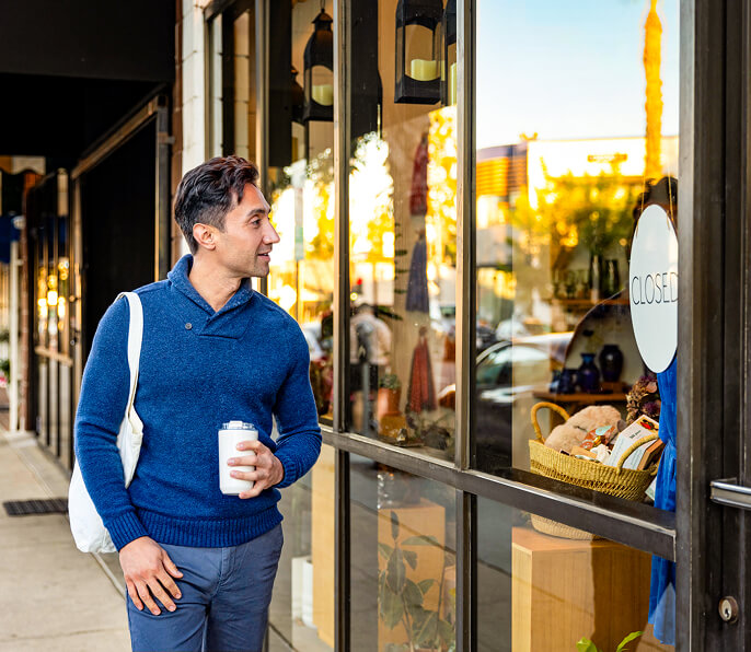 Un hombre con una taza de café y una bolsa de tela mira el escaparate de una tienda que muestra un letrero de "Cerrado" en el distrito comercial del oeste de Salt Lake.