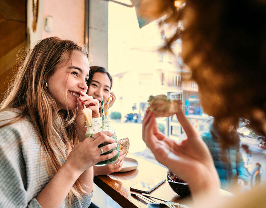 Dos mujeres sentadas en la mesa de un café a lo largo del río Jordan en North Temple, sonriendo y bebiendo de jarras de conserva, mientras una tercera persona sostiene comida en primer plano, capturando el espíritu vibrante de este desarrollo de uso mixto totalmente eléctrico en Utah.