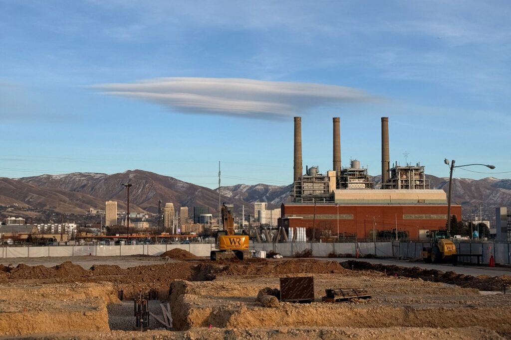 Una obra en construcción para un desarrollo de uso mixto totalmente eléctrico se encuentra frente a un edificio industrial con cuatro chimeneas; edificios de la ciudad y montañas se elevan al fondo bajo un cielo cubierto de nubes.