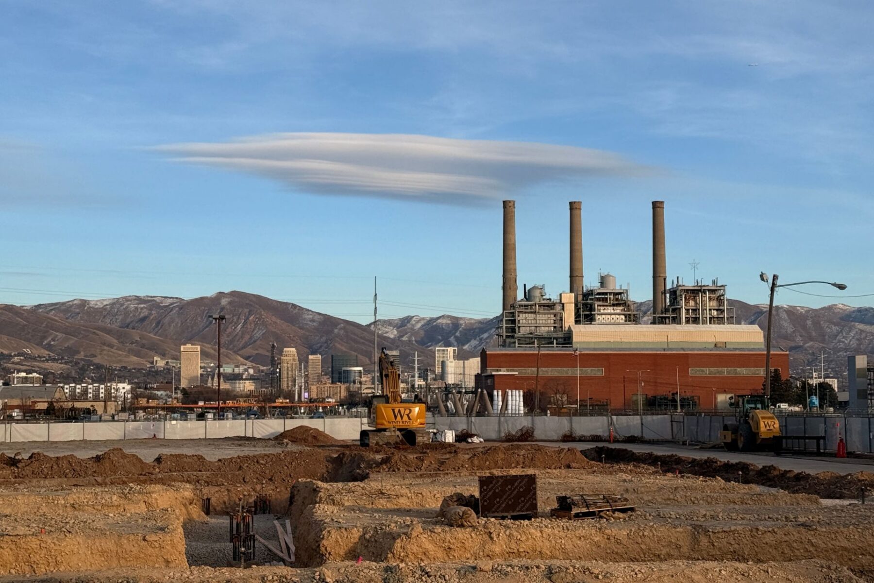 Una obra en construcción para un desarrollo de uso mixto totalmente eléctrico se encuentra frente a un edificio industrial con cuatro chimeneas; edificios de la ciudad y montañas se elevan al fondo bajo un cielo cubierto de nubes.