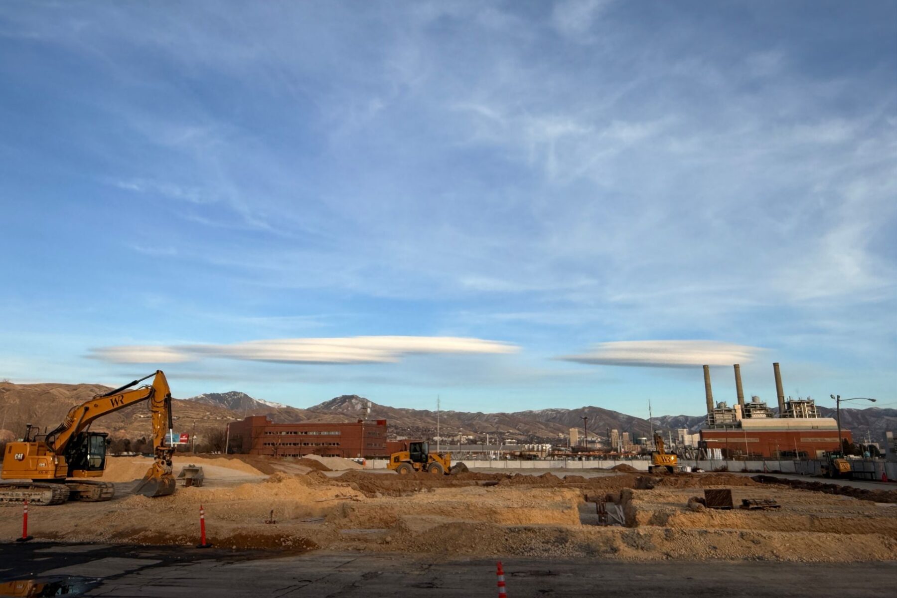 Sitio de construcción con excavadoras y equipo en primer plano, ubicado en el distrito de negocios del oeste de Salt Lake, edificios industriales y montañas al fondo, bajo un cielo azul con nubes lenticulares.