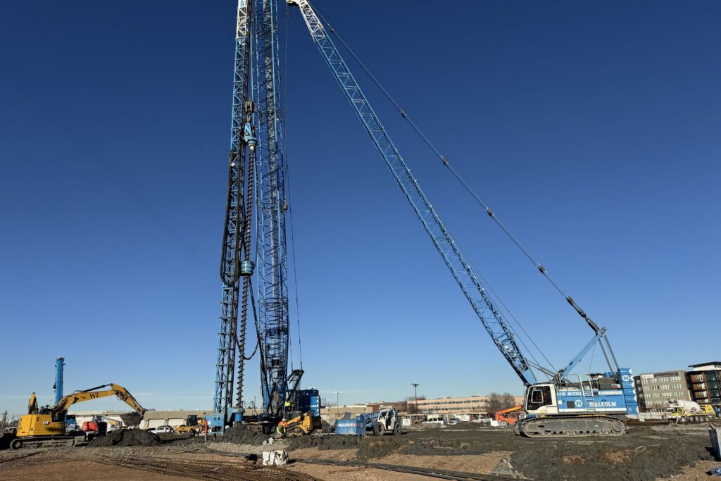 Sitio de construcción con grúas grandes, excavadoras y equipo de perforación operando en un día despejado cerca del distrito de negocios del lado oeste del Lago Salado; edificios visibles al fondo.