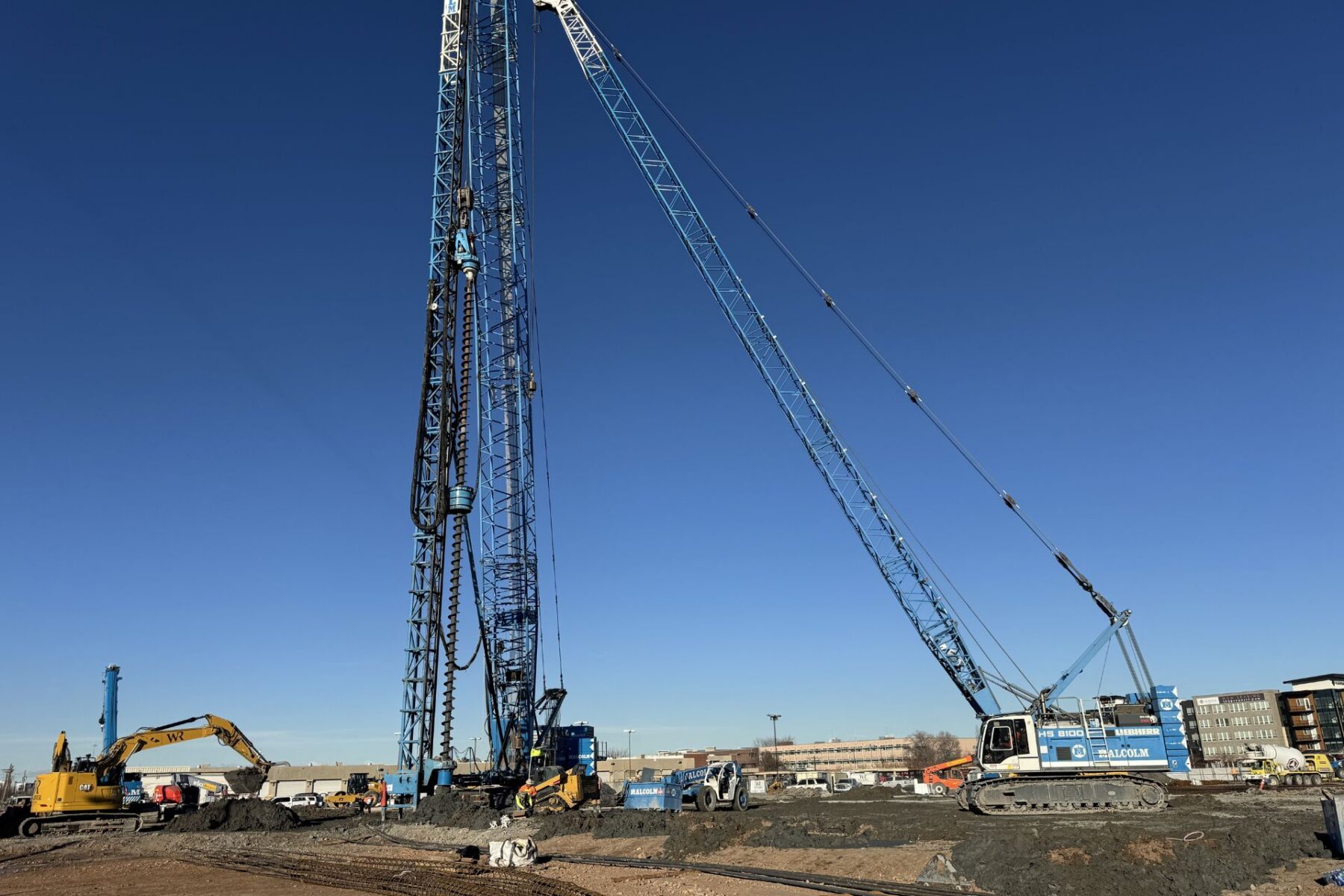 Sitio de construcción con grúas grandes, excavadoras y equipo de perforación operando en un día despejado cerca del distrito de negocios del lado oeste del Lago Salado; edificios visibles al fondo.