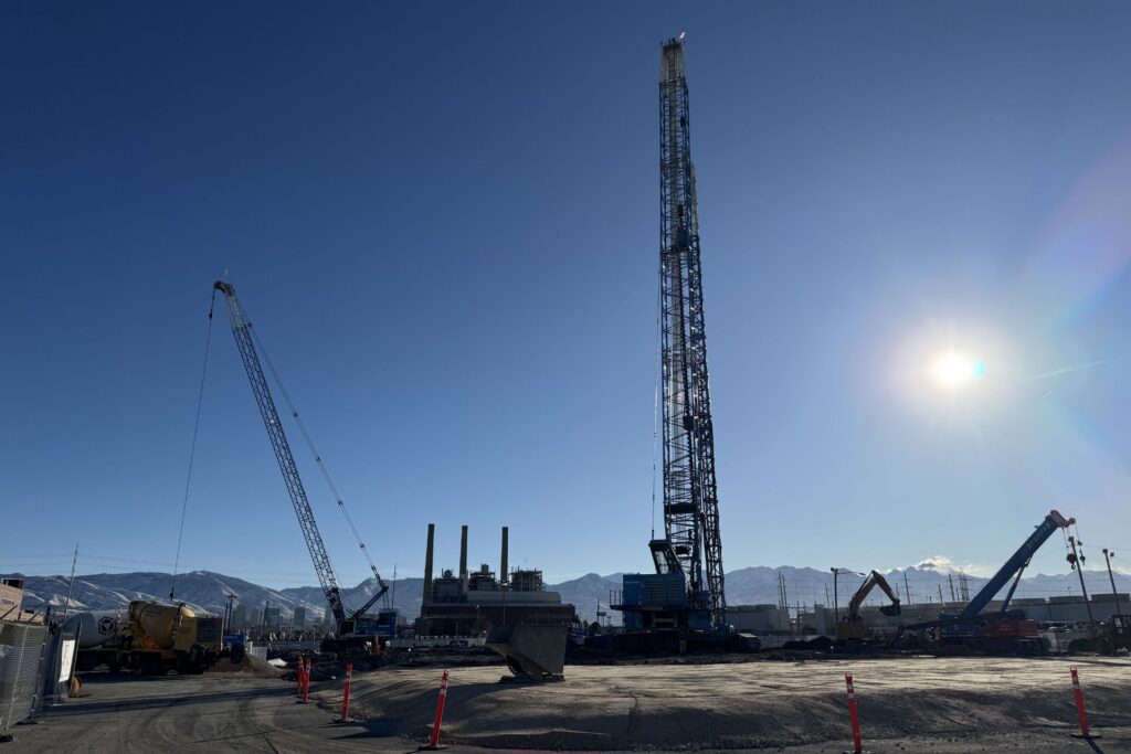 Obra en construcción con grúas, una plataforma de perforación y equipo bajo un cielo despejado cerca del Río Jordán Templo del Norte, con montañas y edificios industriales en el distrito de negocios del oeste de Salt Lake.
