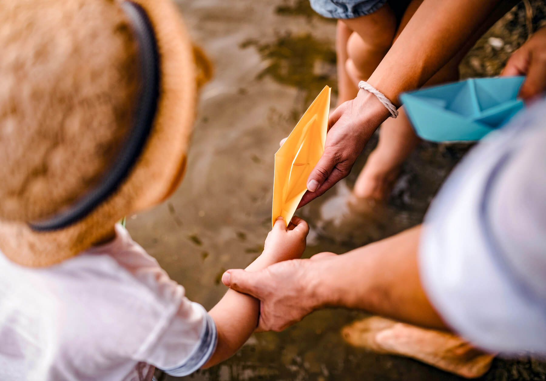 A child in a straw hat holds a yellow paper boat near the North Temple Jordan River, assisted by an adult, with another child and a blue paper boat in the background.