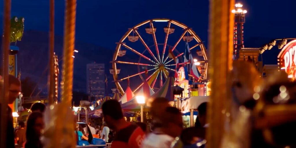 Una rueda de la fortuna intensamente iluminada se alza al fondo de una bulliciosa feria nocturna, ubicada cerca del vibrante distrito comercial Westside de Salt Lake, con multitudes de personas disfrutando del animado ambiente en primer plano.