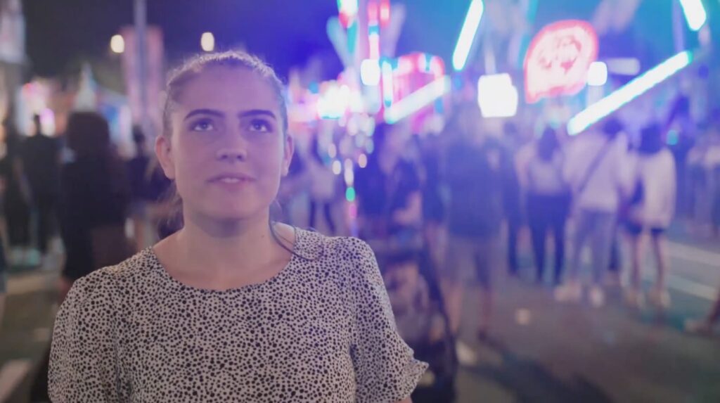 Una mujer con una camisa estampada está al aire libre por la noche con luces de colores y una multitud al fondo, posiblemente en un festival en el distrito comercial del oeste de Salt Lake, conocido por su energía vibrante y su desarrollo mixto totalmente eléctrico en curso.