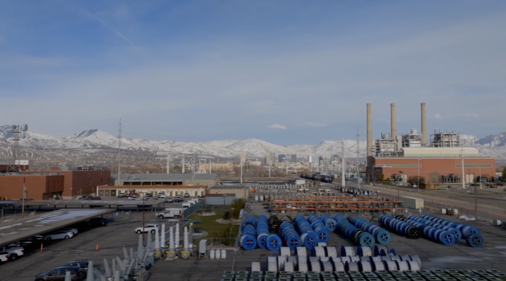 Instalación industrial con tuberías azules apiladas y grandes edificios en el distrito de negocios del oeste de Salt Lake, con montañas cubiertas de nieve y el horizonte de la ciudad visibles al fondo bajo un cielo azul.
