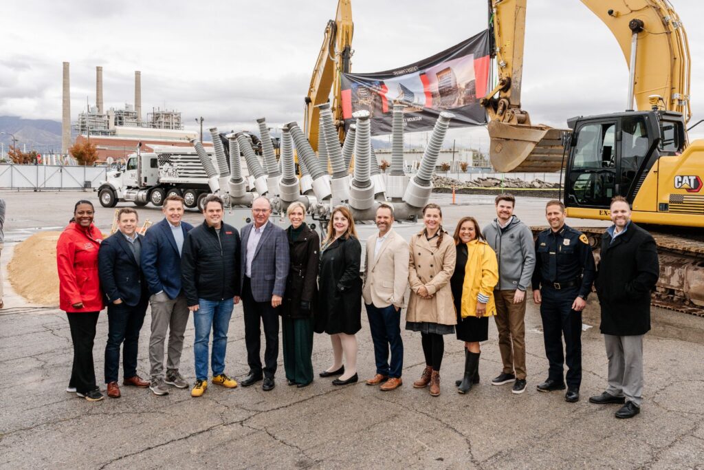 Un grupo de personas está de pie frente a maquinaria de construcción pesada en un sitio industrial al aire libre, posando para una foto en un desarrollo listo para comenzar en Utah.