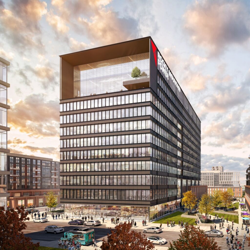 A modern glass office building with a large overhang and rooftop terrace, part of an all-electric mixed-use development, features the "Pratt & Whitney" logo on top. People and cars are visible around the building.