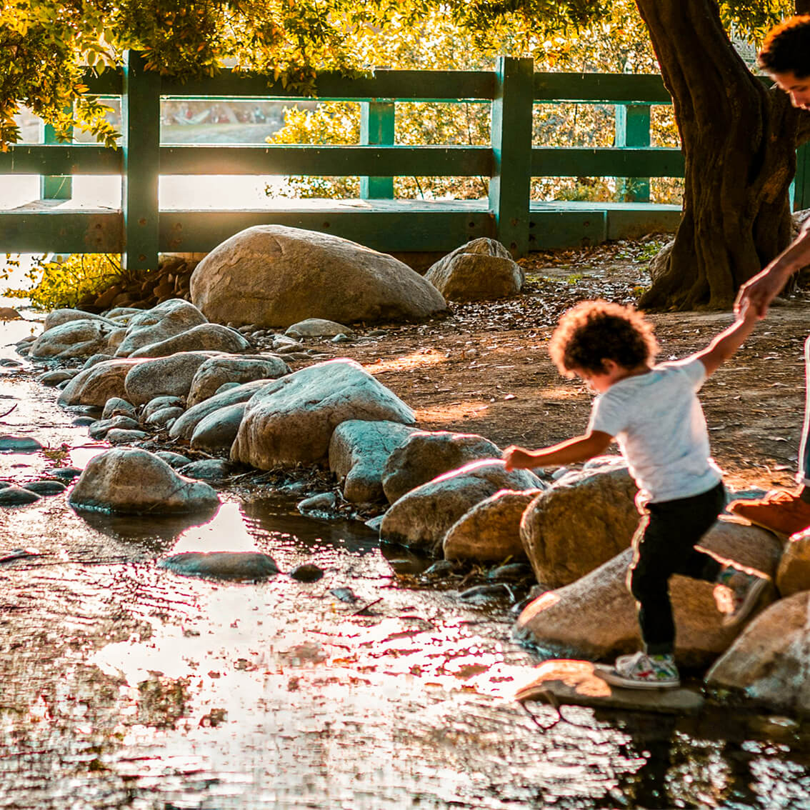 An adult helps a child cross a shallow stream by stepping on rocks in a sunlit park near the North Temple Jordan River, with a green fence and trees framing the vibrant Salt Lake westside business district in the background.