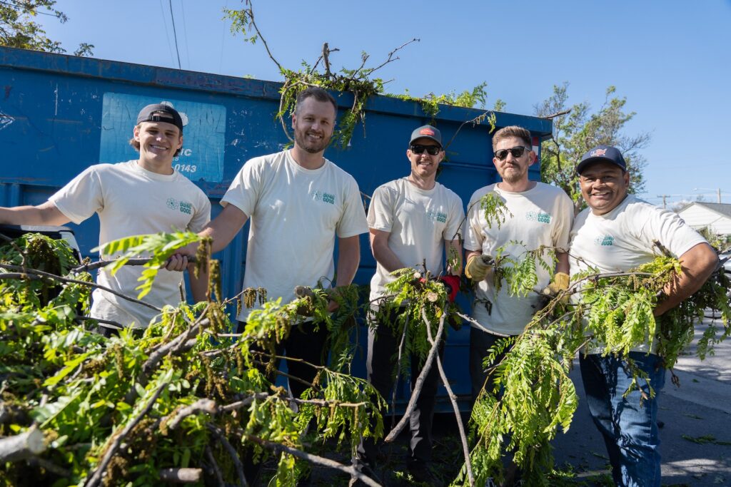 Five people in matching shirts gather tree branches and debris near a large blue dumpster on a sunny day, helping to beautify the Salt Lake westside business district by the North Temple Jordan River.