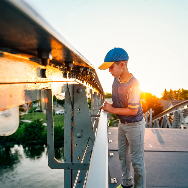A boy wearing a blue and yellow cap stands on a bridge at sunset, looking over the metal railing towards the water below in Salt Lake’s westside business district.