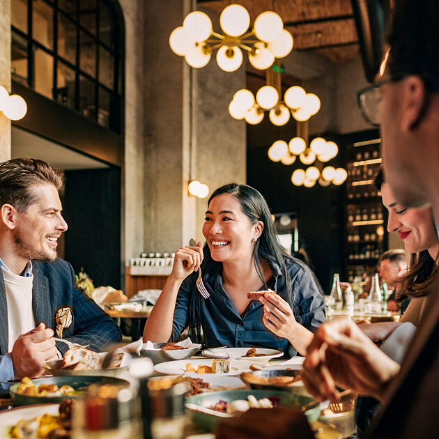 Four people sit at a restaurant table, eating and talking, with modern lighting fixtures and shelves in the background—capturing the vibrant atmosphere of Salt Lake westside business district’s all-electric mixed-use development.