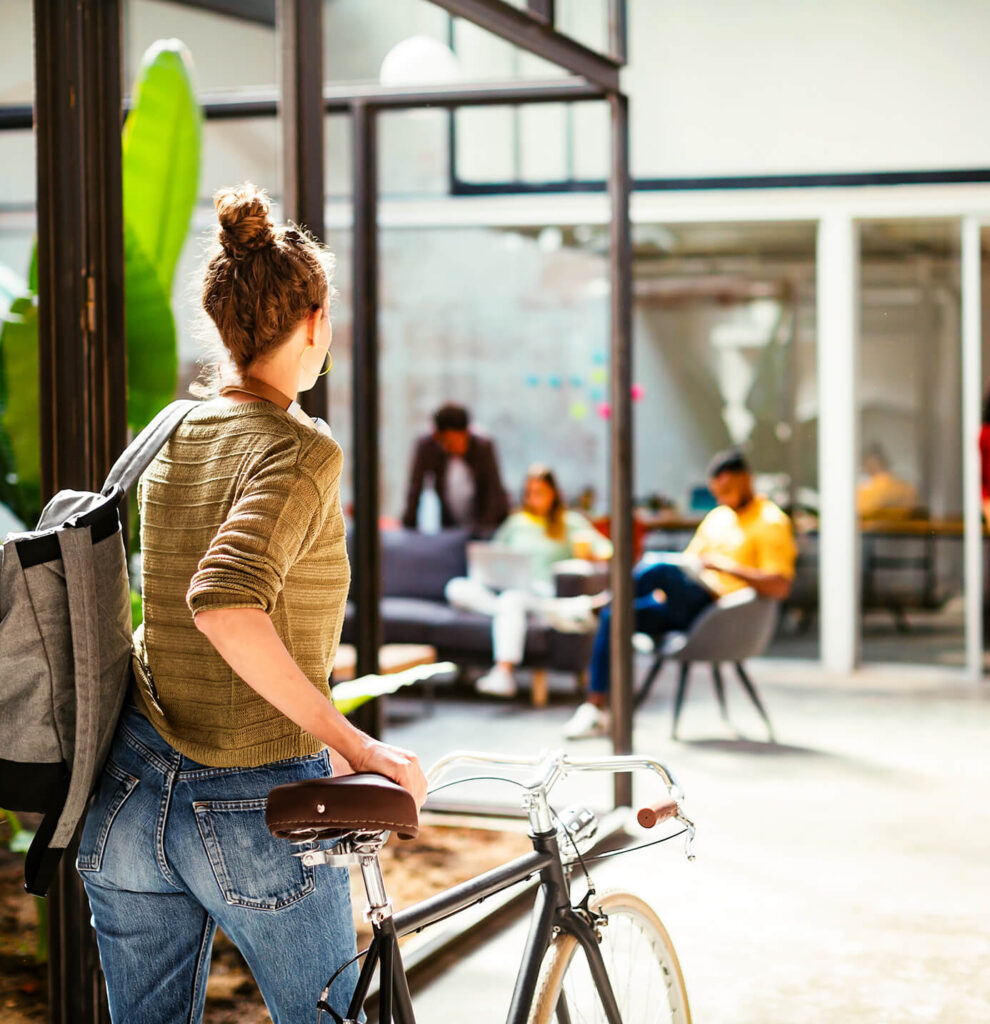 A person stands next to a bicycle, holding the handlebars and wearing a backpack, while looking toward a group of people sitting and talking in an all-electric mixed-use development near North Temple Jordan River.